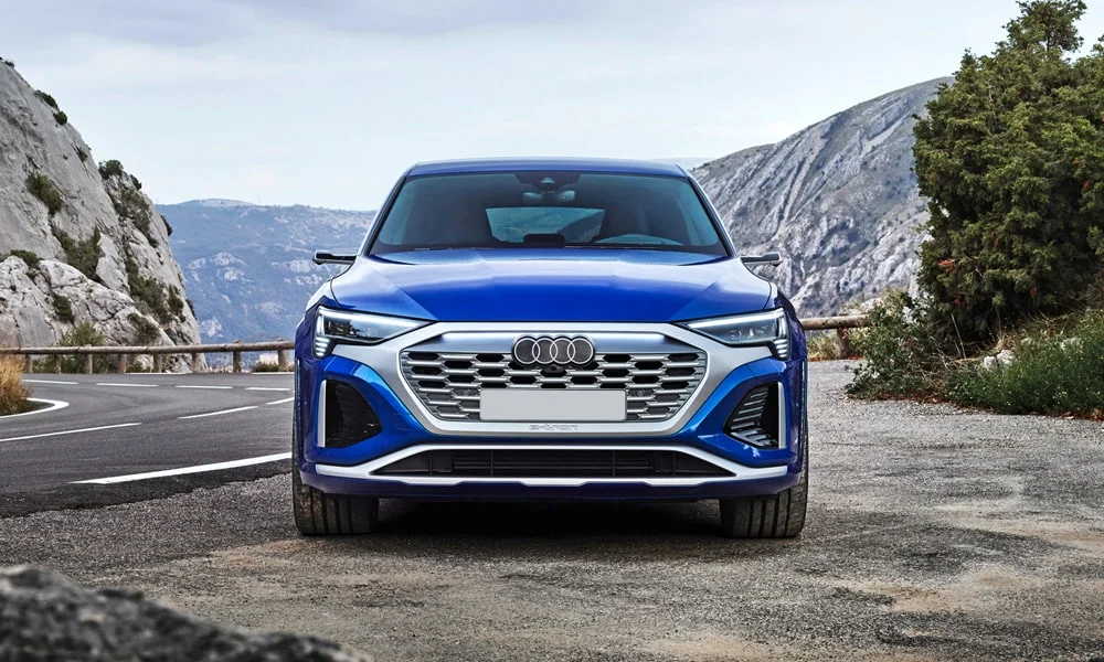 Фото Audi SQ8 Sportback e-tron (2024) — вид спереди, фото 8. Синий кузов, строгая симметрия оптики — базовый ракурс знакомства с дизайном. Рисунок ходовых огней — подпись модели, по нему её узнают в потоке Audi SQ8 Sportback e-tron (2024) — вид спереди, фото 8