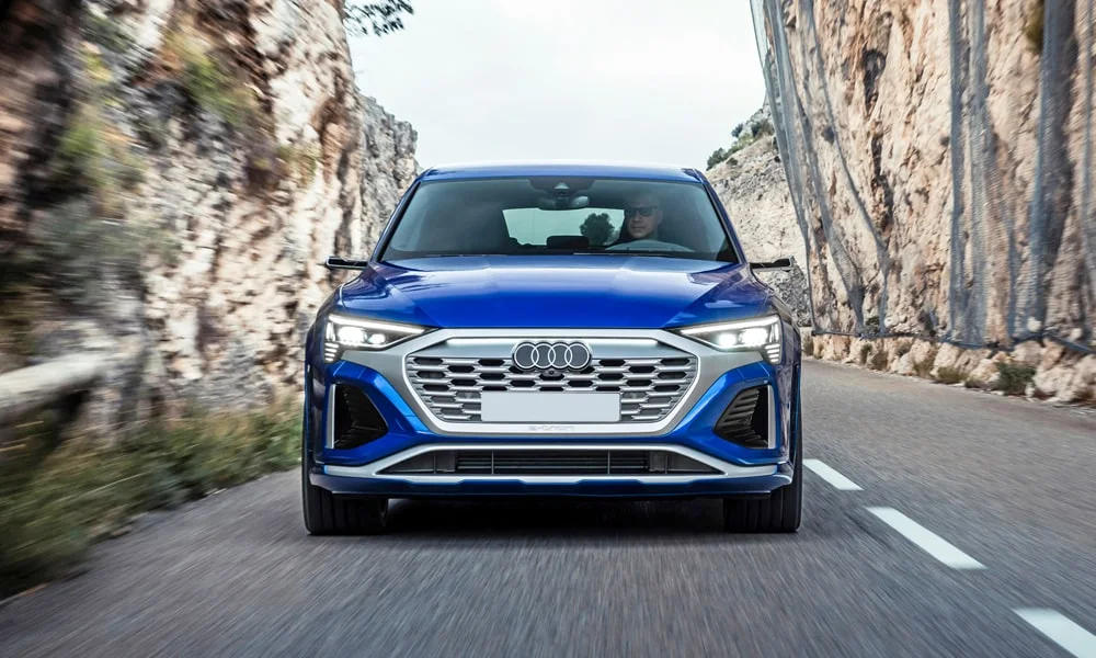 Фото Audi SQ8 Sportback e-tron (2024) — вид спереди, фото 9. Кузов в синем цвете: рисунок оптики и решётки задаёт узнаваемое «лицо» модели. Дневные ходовые огни включаются автоматически с запуском Audi SQ8 Sportback e-tron (2024) — вид спереди, фото 9