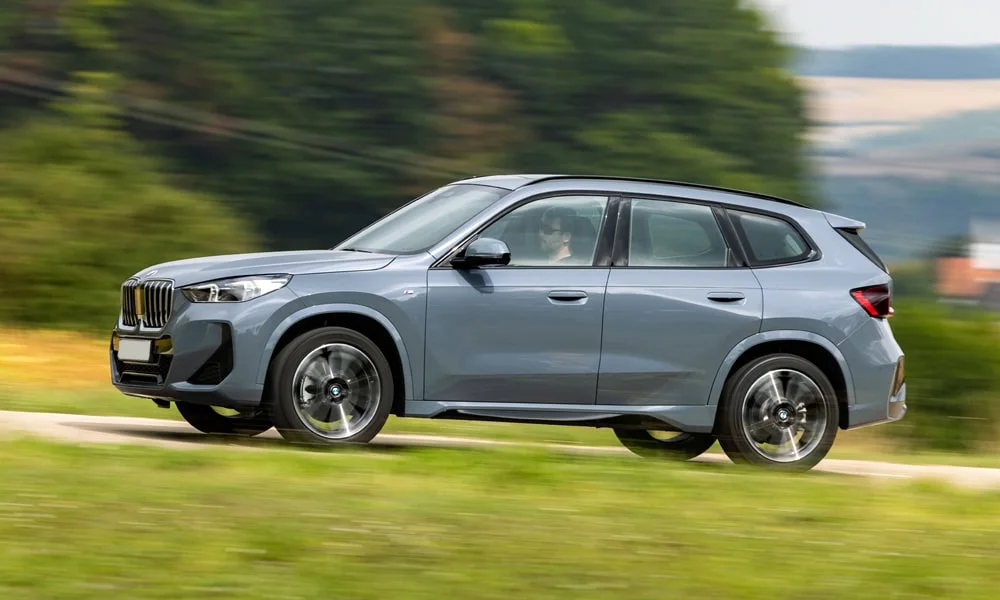 Фото BMW X1 (2024) — вид сбоку, фото 2. Серый кузов в профиль — пропорции, высота посадки и дизайн дисков. Хромированные или чёрные молдинги окон отличают комплектации BMW X1 (2024) — вид сбоку, фото 2