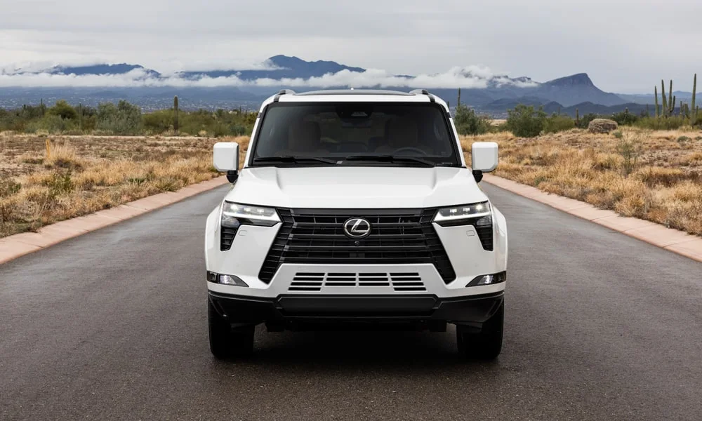 Фото Lexus GX 550 (2024) — вид спереди, фото 7. Белый кузов, строгая симметрия оптики — базовый ракурс знакомства с дизайном. Рисунок ходовых огней — подпись модели, по нему её узнают в потоке Lexus GX 550 (2024) — вид спереди, фото 7