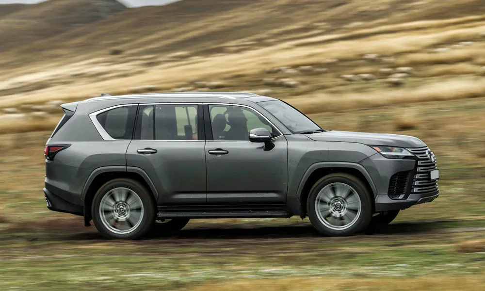 Фото Lexus LX 600 (2022) — вид сбоку, фото 2. Профиль в сером цвете: соотношение колёсной базы и свесов. Хромированные или чёрные молдинги окон отличают комплектации Lexus LX 600 (2022) — вид сбоку, фото 2