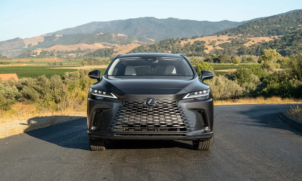 Фото Lexus RX 450h (2024) — вид спереди, фото 7. Кузов в чёрном цвете: рисунок оптики и решётки задаёт узнаваемое «лицо» модели. Рисунок ходовых огней — подпись модели, по нему её узнают в потоке Lexus RX 450h (2024) — вид спереди, фото 7