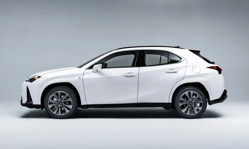 Фото Lexus UX 200 (2024) — вид сбоку. Сбоку читаются линия остекления и характер выштамповок на белом кузове. Хромированные или чёрные молдинги окон отличают комплектации Lexus UX 200 (2024) — вид сбоку