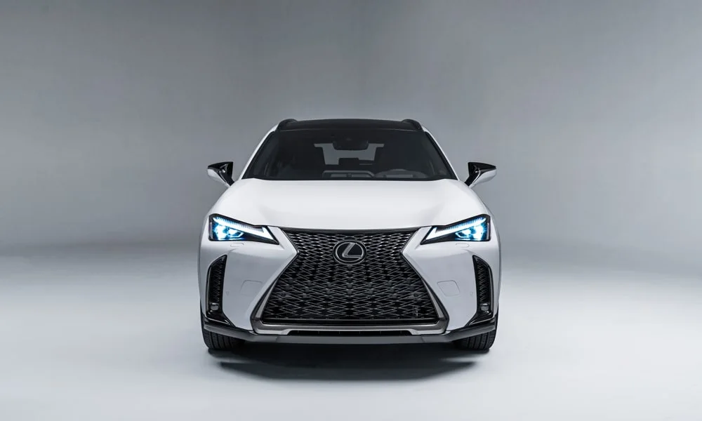Фото Lexus UX 250h (2024) — вид спереди, фото 4. Белый кузов, строгая симметрия оптики — базовый ракурс знакомства с дизайном. Дневные ходовые огни включаются автоматически с запуском Lexus UX 250h (2024) — вид спереди, фото 4