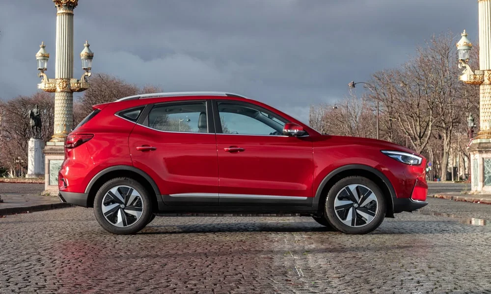 Фото MG ZS EV (2024) — вид сбоку, фото 5. Сбоку читаются линия остекления и характер выштамповок на красном кузове MG ZS EV (2024) — вид сбоку, фото 5