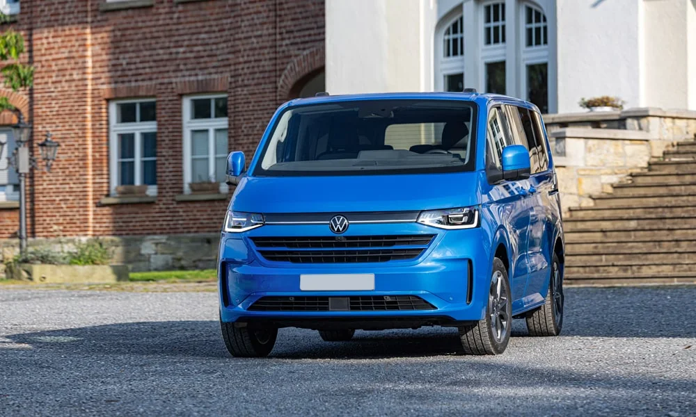 Фото Volkswagen Caravelle (2024) — вид спереди, фото 13. Кузов в синем цвете: рисунок оптики и решётки задаёт узнаваемое «лицо» модели. Рисунок ходовых огней — подпись модели, по нему её узнают в потоке Volkswagen Caravelle (2024) — вид спереди, фото 13