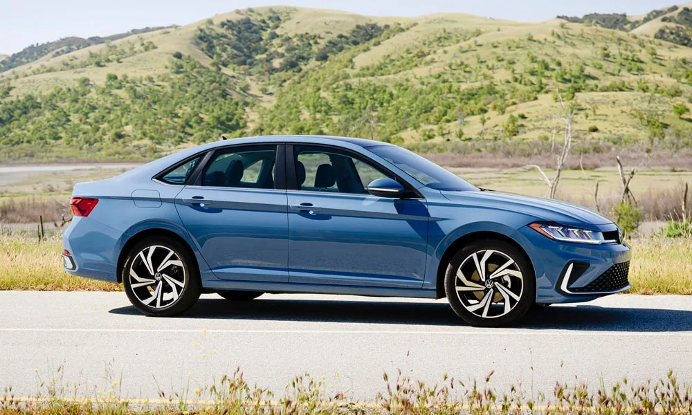 Фото Volkswagen Jetta (2024) — вид сбоку, фото 2. Синий кузов в профиль — пропорции, высота посадки и дизайн дисков. Хромированные или чёрные молдинги окон отличают комплектации Volkswagen Jetta (2024) — вид сбоку, фото 2