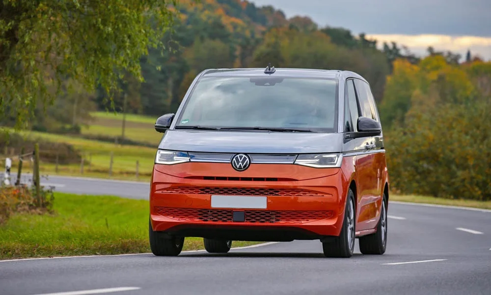 Фото Volkswagen Multivan (2024) — вид спереди, фото 4. Кузов в красном цвете: рисунок оптики и решётки задаёт узнаваемое «лицо» модели. Рисунок ходовых огней — подпись модели, по нему её узнают в потоке Volkswagen Multivan (2024) — вид спереди, фото 4