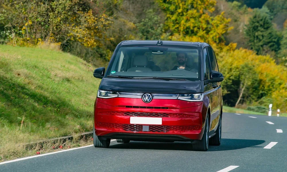 Фото Volkswagen Multivan (2024) — вид спереди, фото 11. Передок в красном исполнении — крупная светотехника и выразительный бампер. Рисунок ходовых огней — подпись модели, по нему её узнают в потоке Volkswagen Multivan (2024) — вид спереди, фото 11