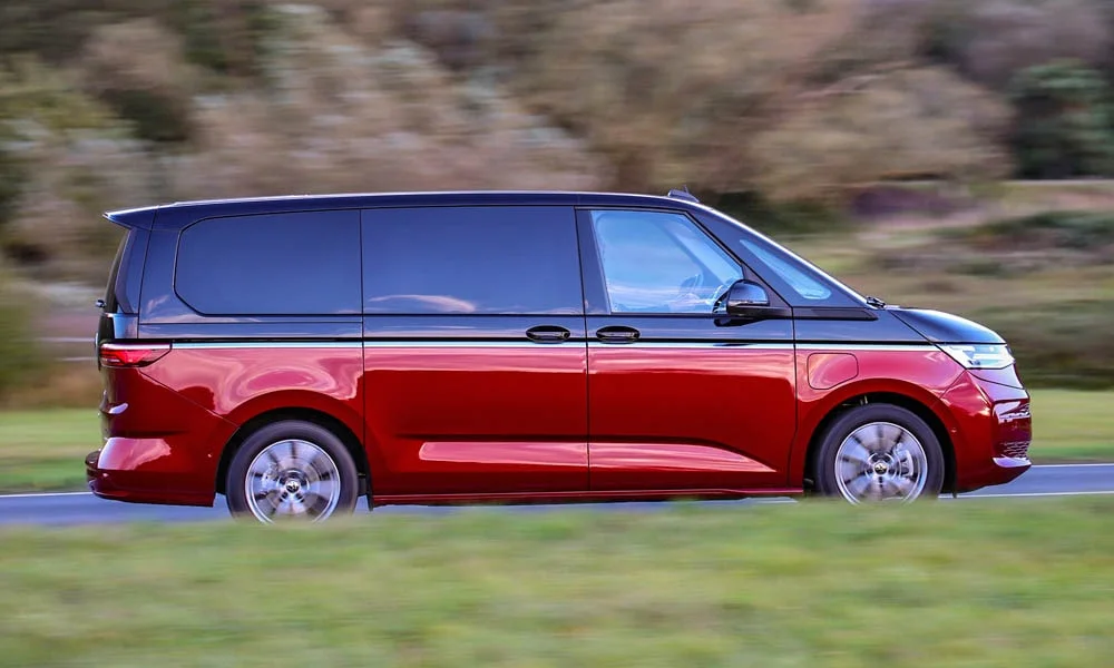 Фото Volkswagen Multivan (2024) — вид сбоку, фото 2. Сбоку читаются линия остекления и характер выштамповок на красном кузове. Хромированные или чёрные молдинги окон отличают комплектации Volkswagen Multivan (2024) — вид сбоку, фото 2